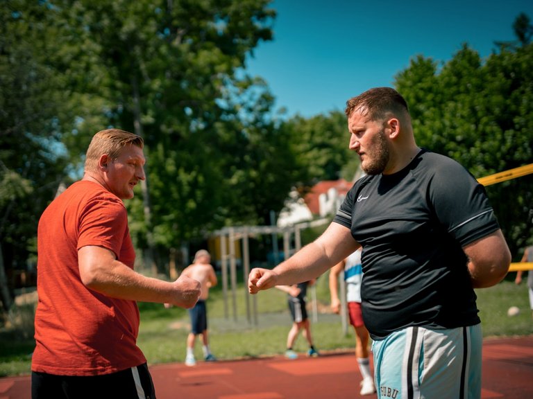 Zwei Männer stehen sich auf einem Sportplatz gegenüber. Der Mann im orangefarbenen T-Shirt und der Mann im schwarzen T-Shirt heben ihre Fäuste, als wollten sie sich die Fäuste stoßen. Im Hintergrund sind unscharf weitere Personen und Bäume zu erkennen.