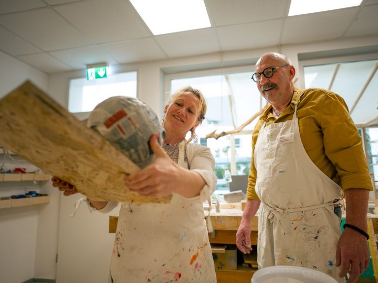 Zwei Künstler in schmutzigen weißen Kitteln stehen in einer Werkstatt. Die Frau hält ein großes Kunstobjekt aus Holz und Papiermache hoch und lächelt, während der Mann mit Brille und Schnurrbart ihr zufrieden zuschaut.