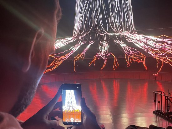 Ein Mann fotografiert mit seinem Smartphone eine Lichtinstallation. Im Hintergrund ist ein beleuchteter Baumstamm mit weit verzweigten, leuchtenden Wurzeln in Weiß und Orange zu sehen, deren Spiegelung sich auf dem glatten Boden widerspiegelt.